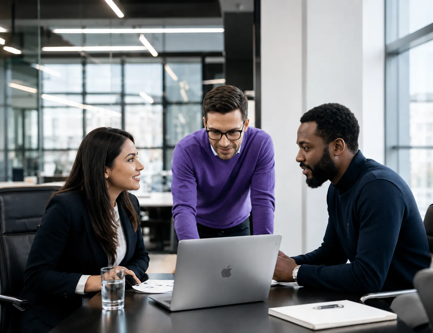 Three colleagues reviewing work together at a laptop in a glass-walled office